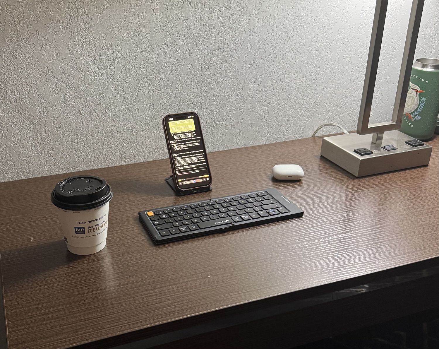 Mobile vibe coding setup in a hotel room: iPhone on stand with foldable keyboard, coffee, and AirPods